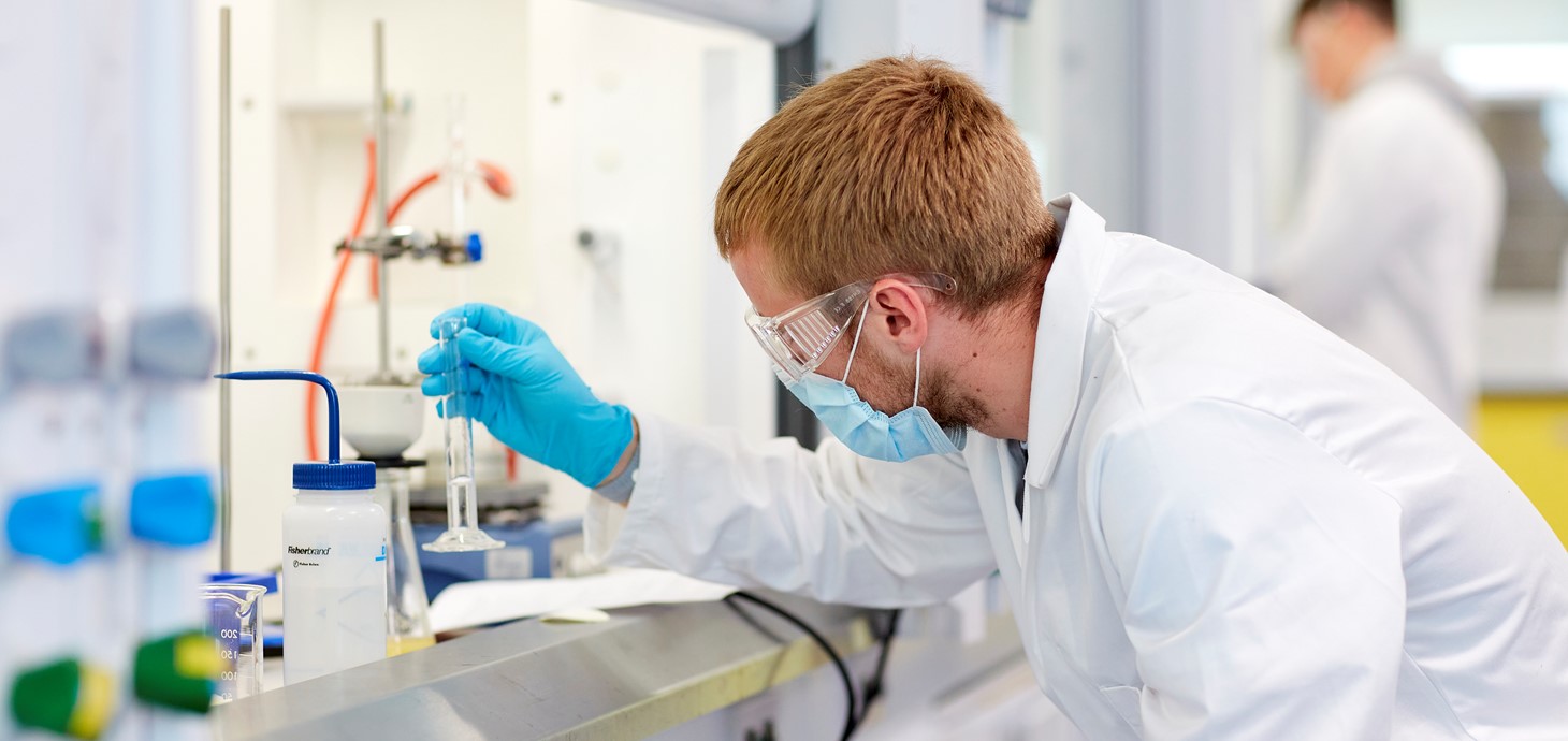 A person wears a surgical mask, white lab coat and gloves as they work in a chemistry lab.