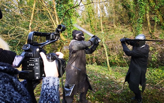 Two men dressed as medieval knights in armour fighting outdoors  with swords while being photographed by a person with a camera.

