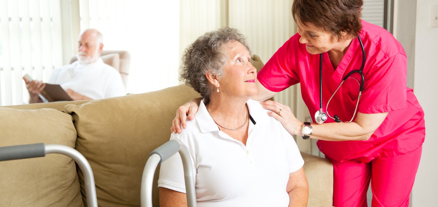 Older woman sitting down looking up at a nurse in uniform who has her arms on her shoulders and smiling