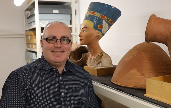 Man smiling inside a storeroom surrounded by shelves holding a variety of Egyptian artefacts.
