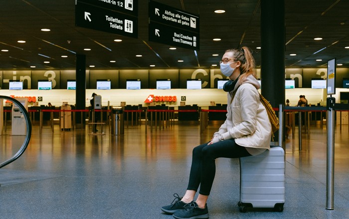Young woman wearing a face mask sitting on a suitcase in an airport terminal