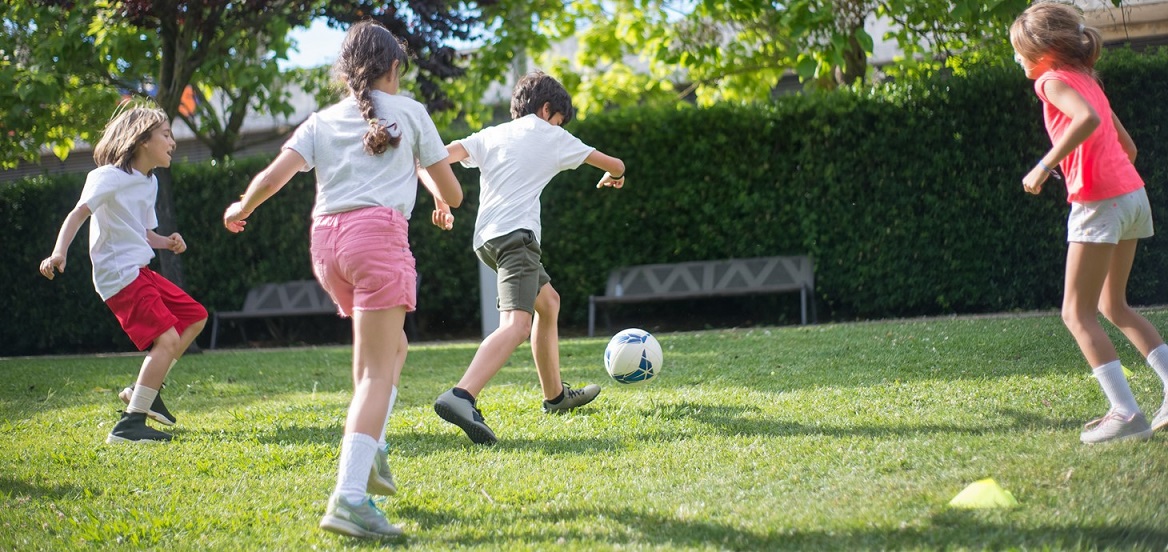 Four children are playing football in a park in front of a hedge and two benches.