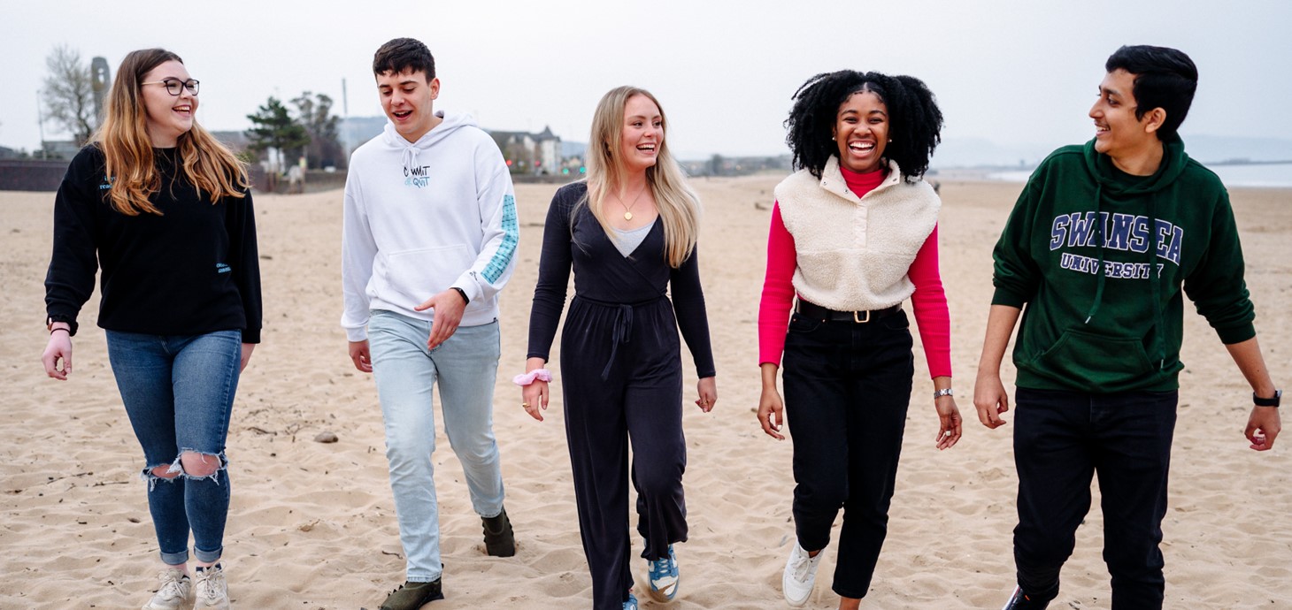 A group of five male and female students are laughing together as they walk on a beach. 