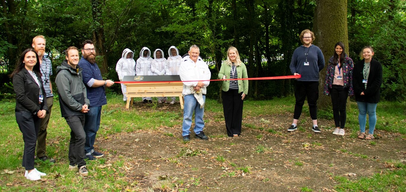 The official beehive opening ceremony on Singleton Campus. Founder of Bee1, Mark Douglas (Left) and Lucy Griffiths (right), Head of SEA, cut the ribbon, as beekeeping volunteers and staff members look on.