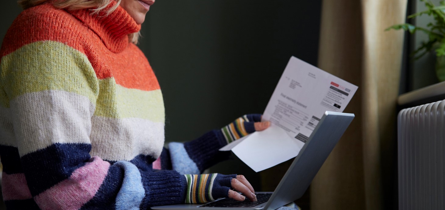 A woman wrapped in a blanket and wearing a thick jumper and gloves looks at a laptop while holding a utility bill in her hand.