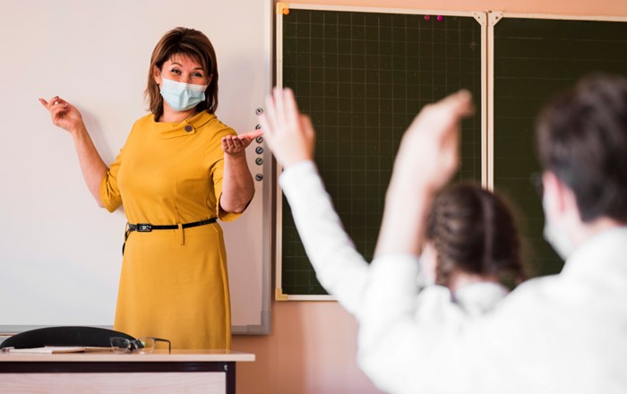 Female teacher wearing a facemask standing at a white board at the front of pupils.