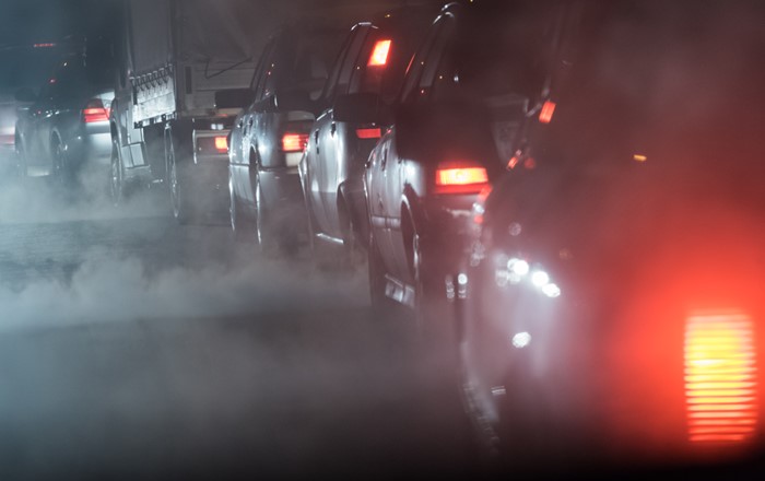 A row of cars in a traffic jam at night with exhaust fumes rising in the air surrounding them. 
