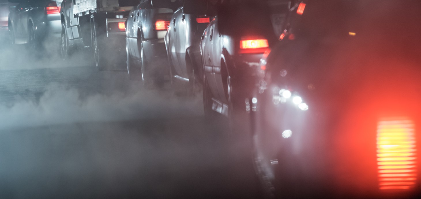 A row of cars in a traffic jam at night with exhaust fumes rising in the air surrounding them. 