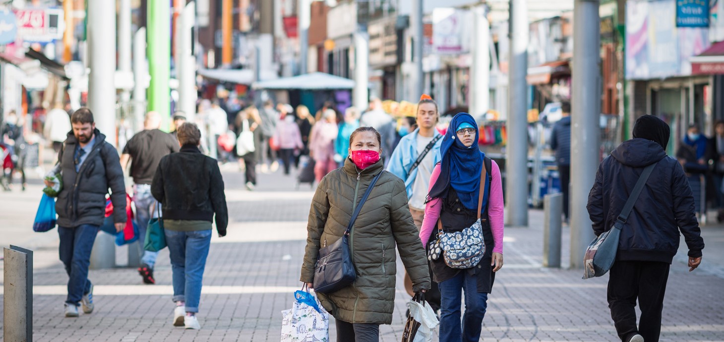 Woman wearing a facemask front and centre walking along a pedestrianised shopping street with other people in background 