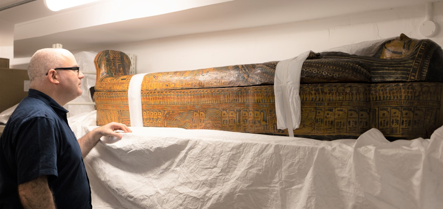 Man standing in storeroom looking at a wooden Egyptian coffin.