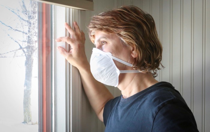 Woman wearing a mask looks sadly out the window of her home.