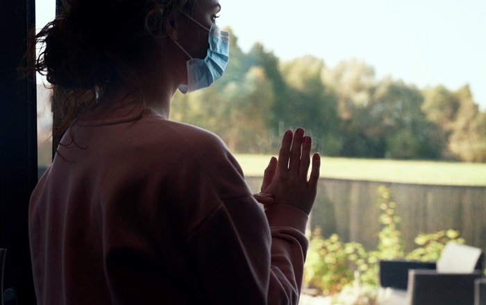 Back view of a woman with protective mask standing indoors looking out of a window.