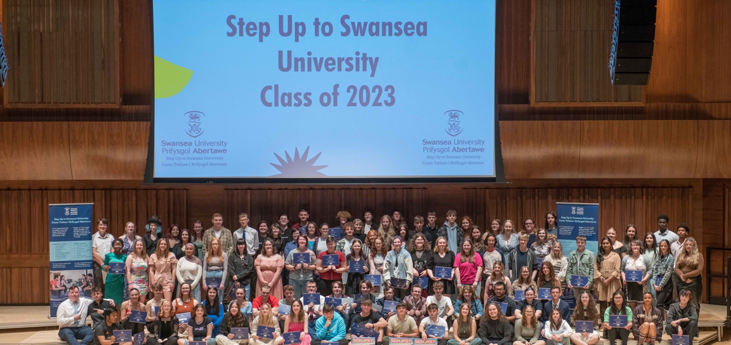 A large group of young people are pictured in a hall in front of a backdrop with the words ‘Step Up to Swansea University Class of 2023’ on it.
