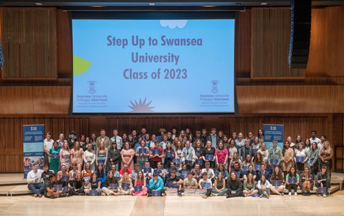 A large group of young people are pictured in a hall in front of a backdrop with the words ‘Step Up to Swansea University Class of 2023’ on it.