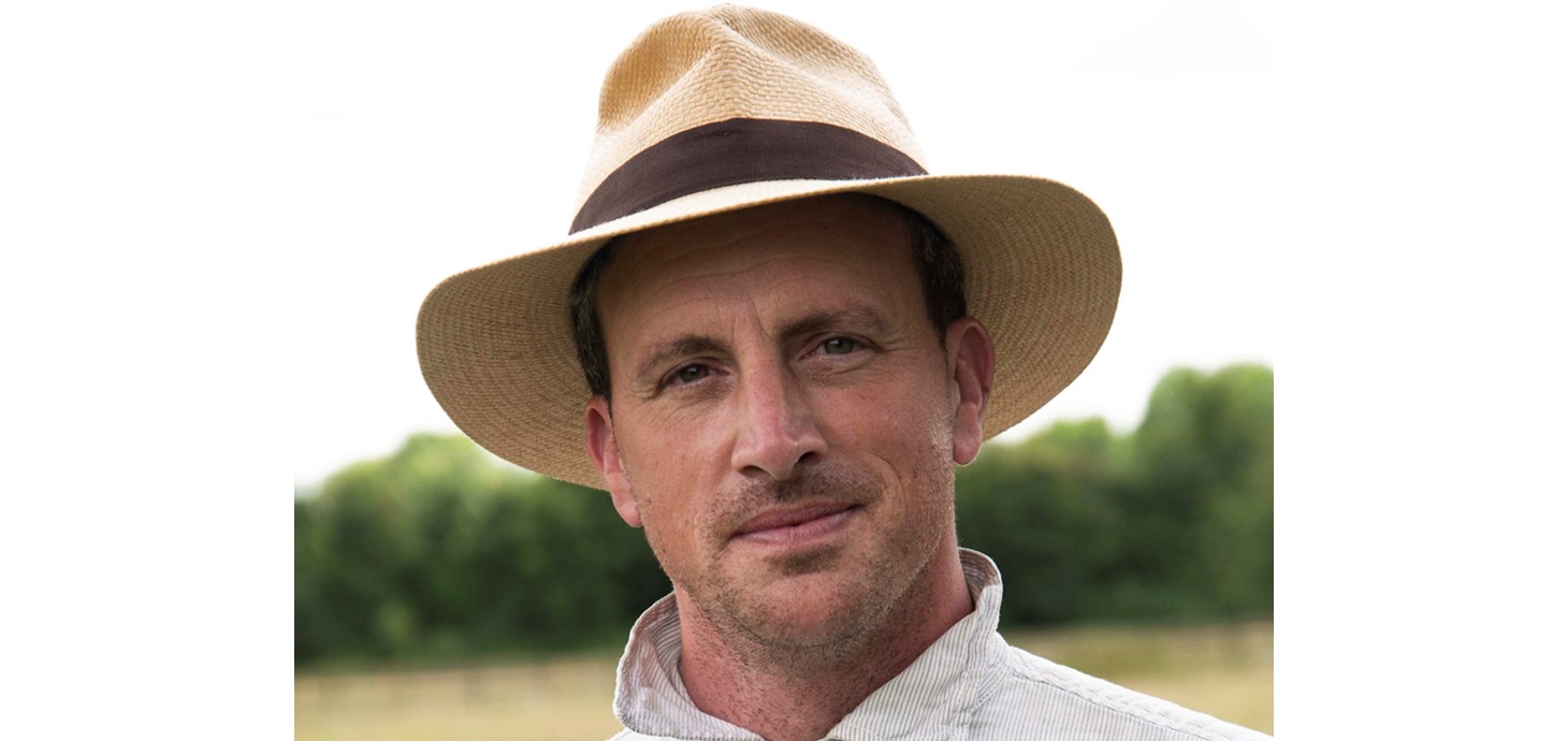 Dr Alexander Langlands is shown in a head and shoulders picture wearing a white shirt with thin blue stripes and a straw hat with a brown band around it.