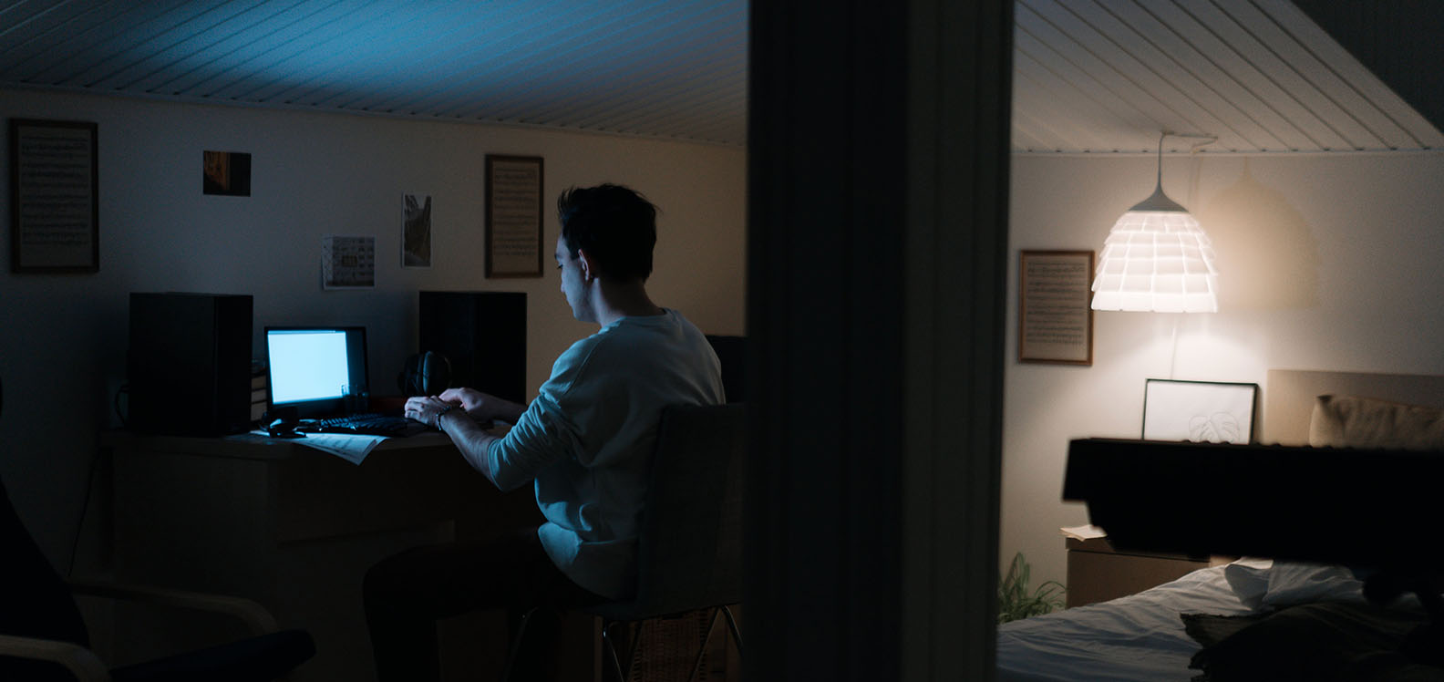 A young man alone in his darkened bedroom sat at a desk looking at his laptop