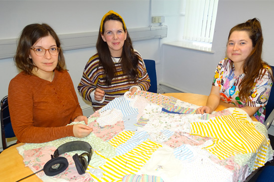 Three seated women sewing a large quilt which laid out on the table in front of them.