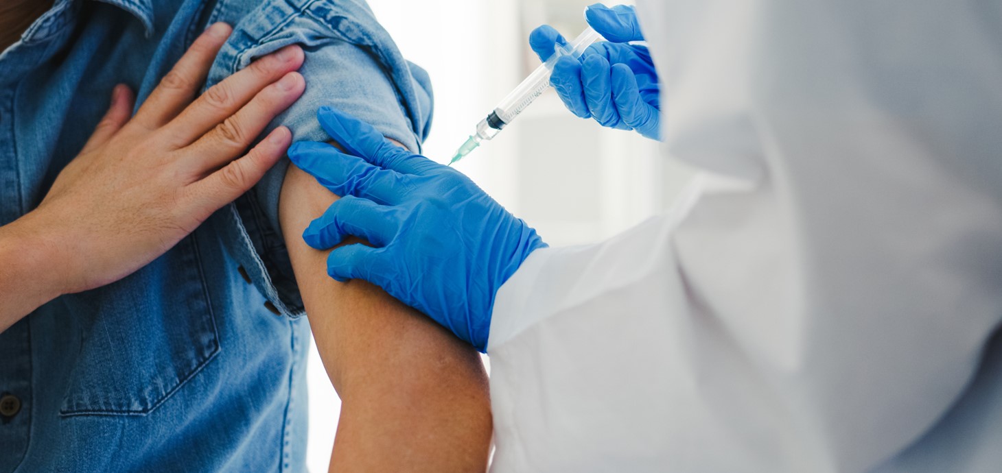 Person in white coat wearing gloves giving an injection into the left arm of another person