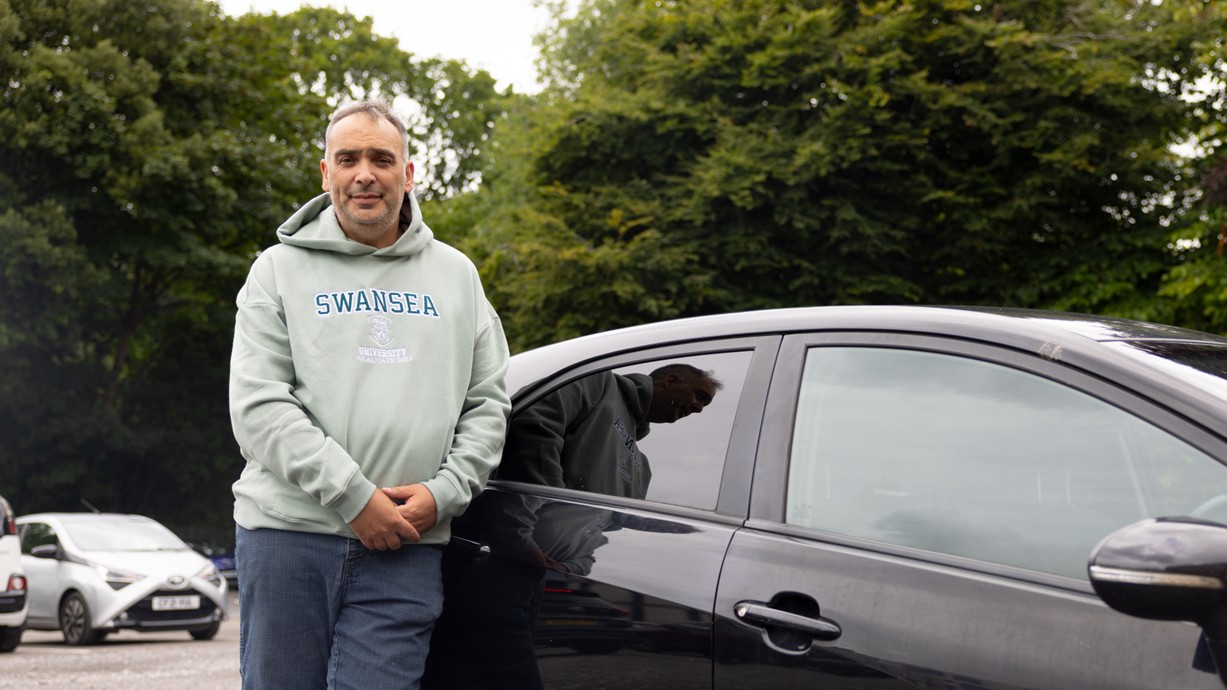 Mark wears a grey Swansea University hoodie as he learns against a black car.