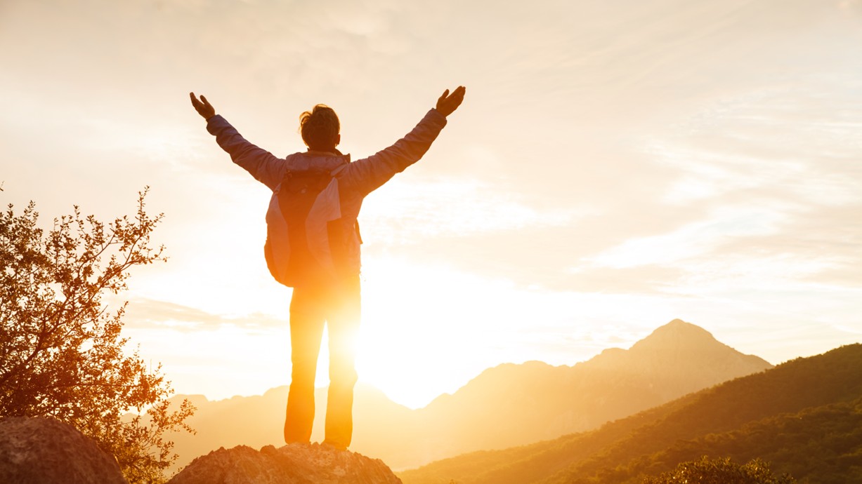 An example of someone thriving: A hiker stands on a mountain cliff with a backpack. The sun rises behind them. Credit: lenina11only | Shutterstock.