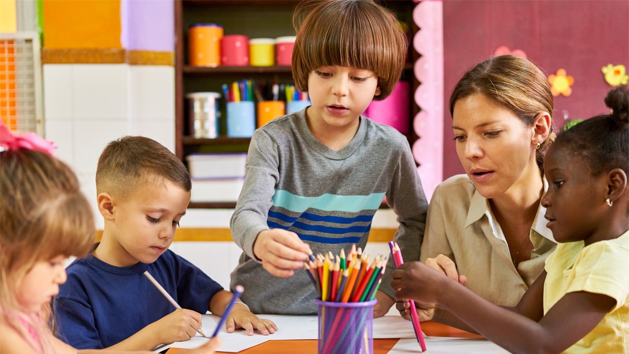 Four young children and one woman sitting round a classroom table using pens and paper