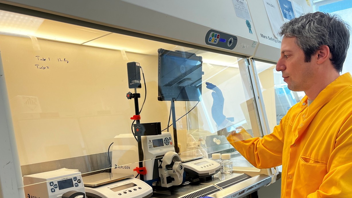 Dr David Howard, Senior Postdoctoral Research Associate, wearing a yellow lab coat and blue gloves, as he operates the Repligen tangential flow filtration system used for exosome purification.