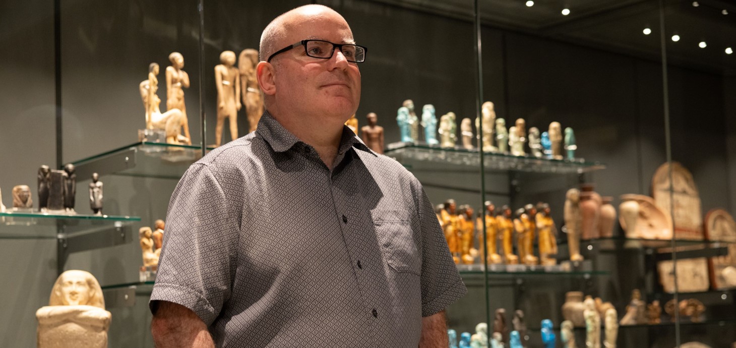 Man standing in front of display cases containing small statuettes.