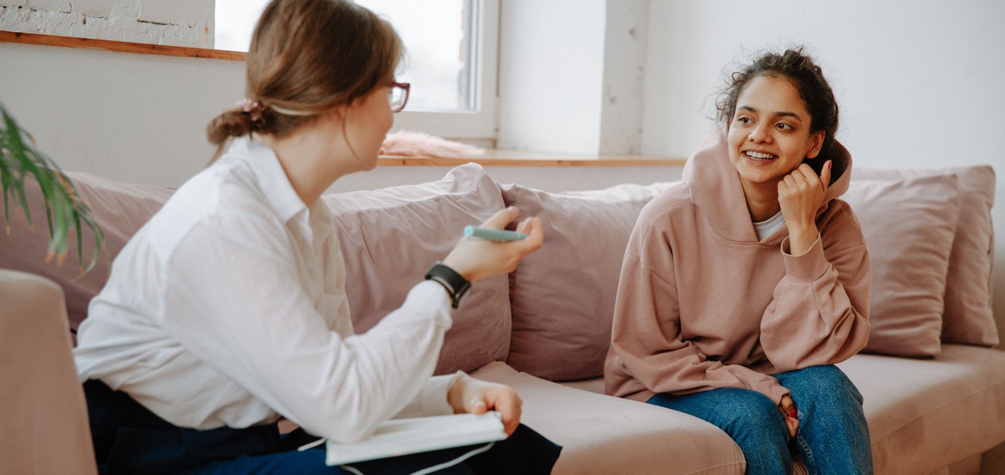 Two women sitting on a sofa talking to each other, one holding a notebook and pen