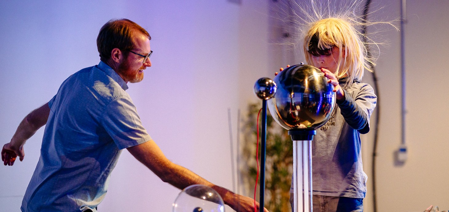 A man and child demonstrate a Van de Graaff generator as the child's hair stands on end, creating a striking, playful static electricity experiment.