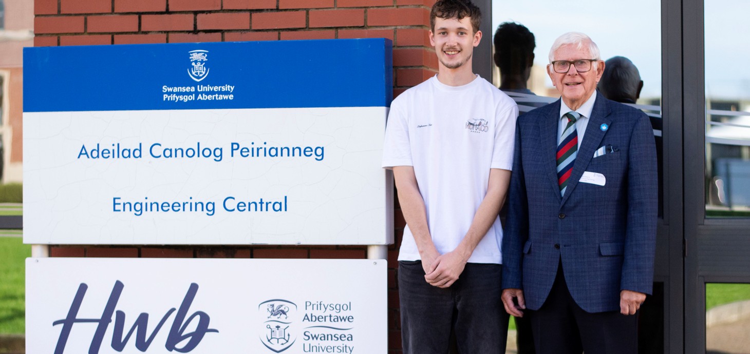 Young adult man and elderly man standing outside a building alongside a sign.