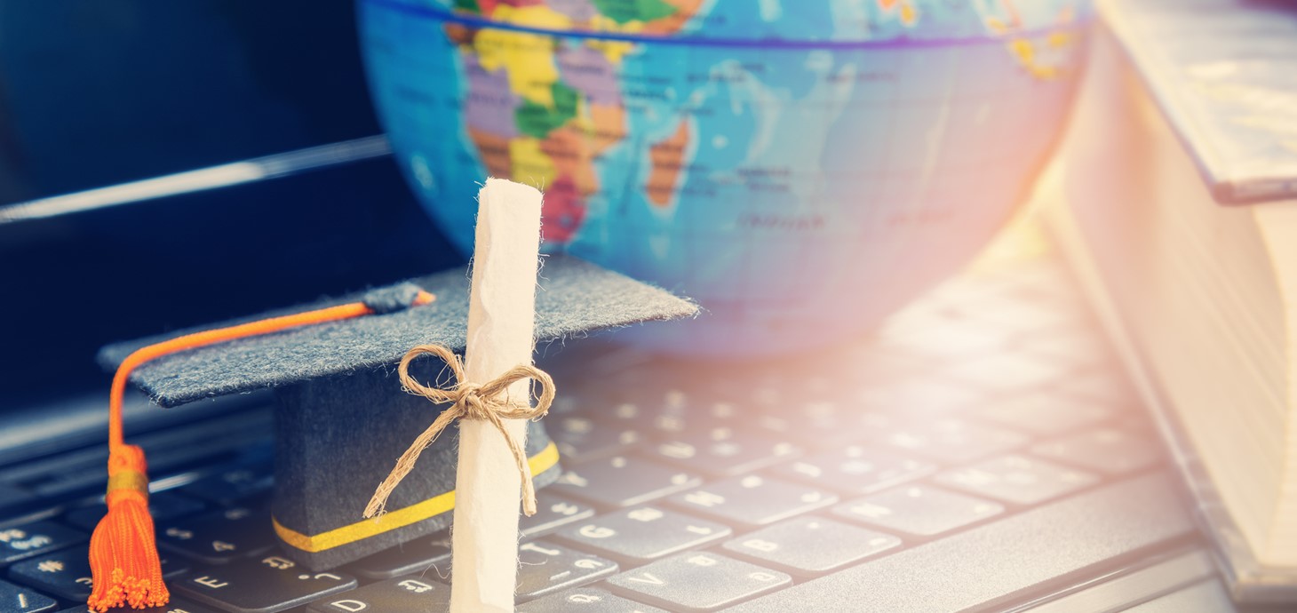 World globe and book in background and a computer keyboard in the foreground with a miniature mortarboard and degree scroll resting on it.