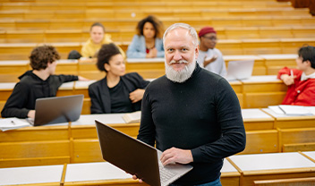 A smiling male lecturer stands in front of his students with a laptop in hand