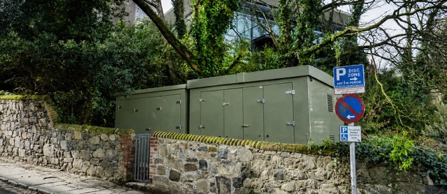 Two large grey fibreglass buildings with double doors sit behind a stone wall, with trees in the background.