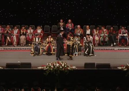 ARCS team member in black robes on stage receiving an award from the Chancellor in gold and black robes during a university graduation ceremony, with faculty in academic robes seated nearby.