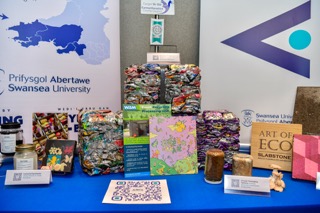 A display table showcasing products from ARCS-supported enterprises, including colourful blocks of crushed aluminium cans, printed building material samples labelled Art of Eco, a glass jar of water treatment beads, cakes, mushrooms and promotional leaflets