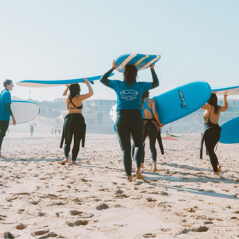 A group of friends walk along the beach in wetsuits, carrying surfboards on their heads 