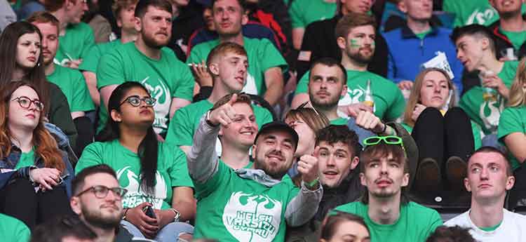 Swansea University students dressed in green t shirts in the stands at the Swansea.com stadium, one gives a thumbs up to the photographer