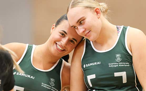Two female students from the netball team smiling, one resting her head on the other's shoulder