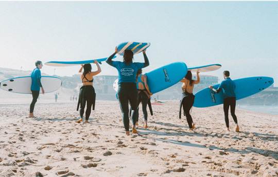 A group of friends walk along the beach wearing wetsuits and carrying surfboards on their heads. 