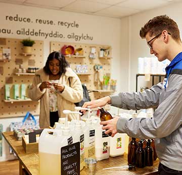 An image of a male and female student in the zero waste shop Root Zero on Singleton Campus. The male student is in the foreground filling a container. The female student is in the background looking at something.