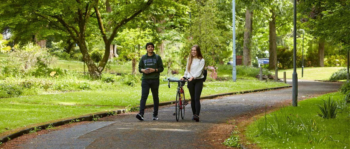 Two students walking through Singleton Campus botanical garden area, with one student pushing a bike.