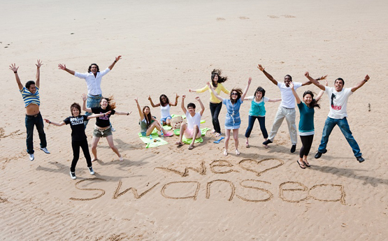 Students jumping in the air on the beach with I heart Swansea drawn in the sand in front of them
