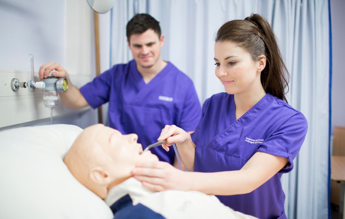 Two students wearing scrubs are practicing on a dummy model patient