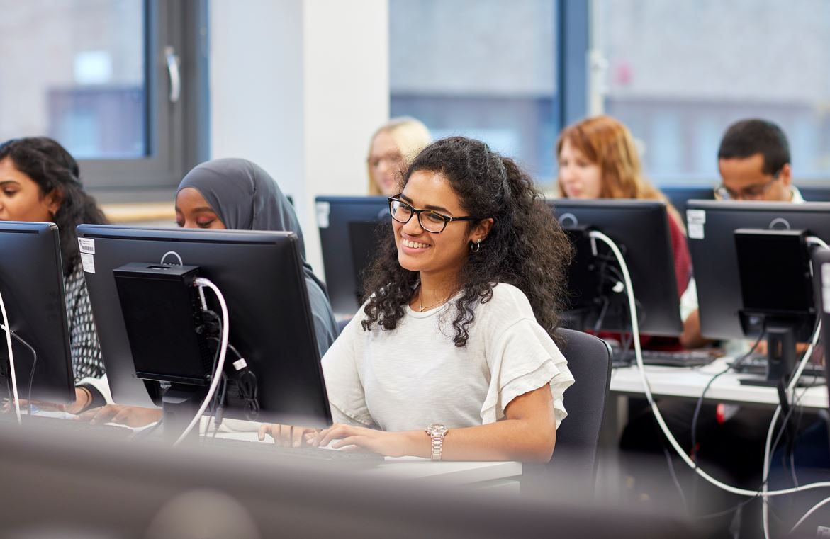 Student sat at computer smiling with other students in background
