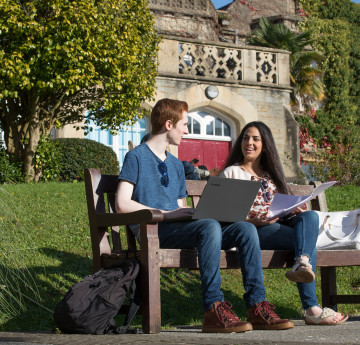 A female and male student sitting on a bench outside with their laptop and papers